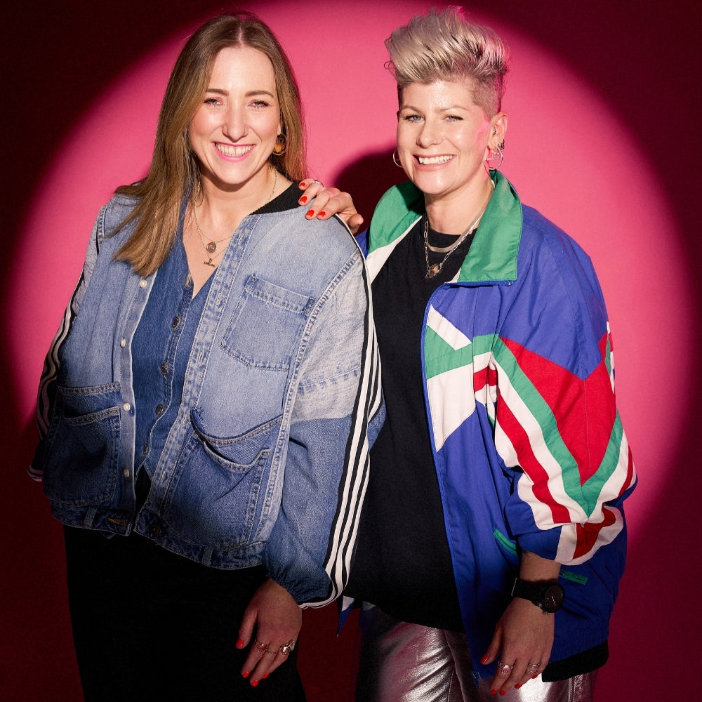 Two women smiling with denim jacket and multicoloured jacket, posing against a pink studio backdrop